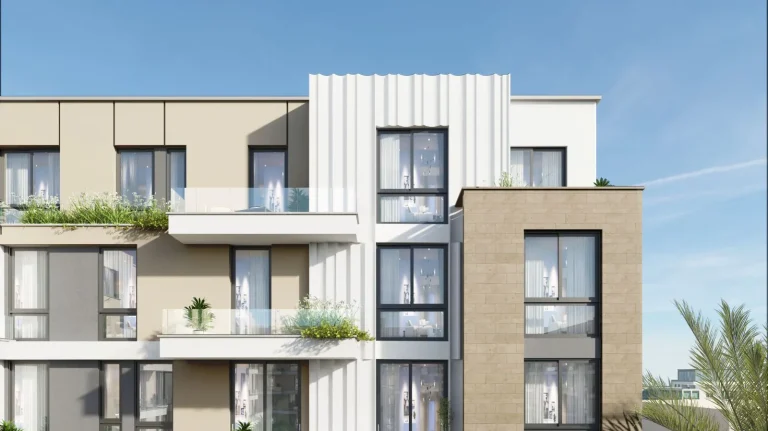 Contemporary multi-story residential building with a beige and white color scheme. The building has large windows and glass-railed balconies with potted plants. The facade features a mix of smooth and textured finishes. Palm trees and other greenery are present, with a clear blue sky in the background