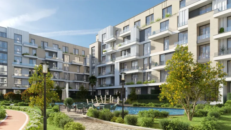 Modern residential apartment complex with multiple contemporary buildings featuring large windows, balconies, and a mix of glass and metal materials. The complex is surrounded by well-maintained landscaping, including trees, shrubs, a small pond, and paved walkways. There are outdoor seating areas with tables, chairs, and street lamps for lighting.