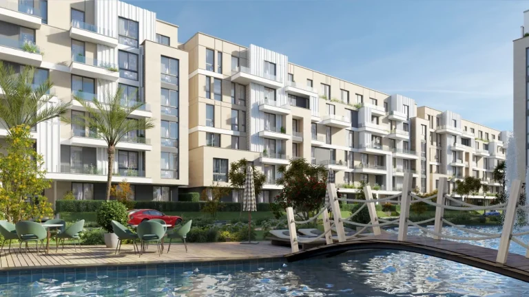 Modern residential complex with multiple apartment buildings featuring large windows, balconies, and a mix of glass, metal, and concrete materials. The complex is surrounded by well-maintained landscaping, including trees, shrubs, and grassy areas. In the foreground, there is a swimming pool with a wooden deck, outdoor seating, and a small bridge crossing over the pool. The scene is set under a clear blue sky.