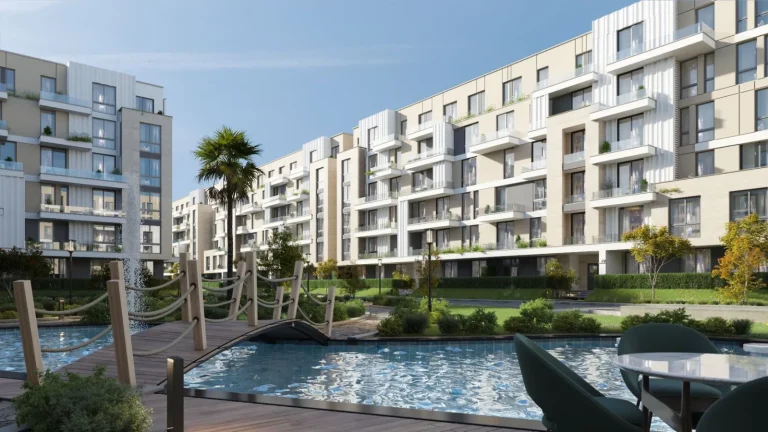 Modern residential complex with mid-rise apartment buildings featuring large windows and balconies. The buildings are surrounded by landscaped gardens with trees and shrubs. A small wooden bridge crosses over a water feature, creating a picturesque outdoor setting with seating areas for residents