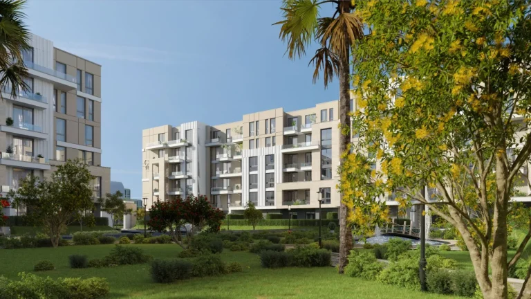 Modern residential complex with multiple mid-rise apartment buildings featuring beige and white facades, large windows, and balconies. A well-maintained garden with palm trees and flowering trees with red and yellow blossoms is in the foreground. A small bridge over a water feature adds to the aesthetic appeal. The sky is clear and blue, suggesting a pleasant day