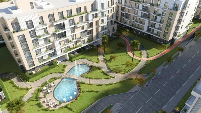 Modern residential complex with multiple apartment buildings surrounding a landscaped courtyard. The courtyard features a uniquely shaped swimming pool with a small bridge crossing over it, surrounded by well-maintained green spaces, walking paths, and various plants and trees. The buildings have multiple floors with balconies, and the overall design is contemporary with a mix of light and dark exterior colors