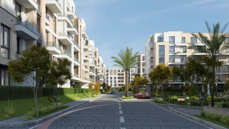 Modern residential area with multi-story apartment buildings on both sides of a wide, paved street. The buildings feature balconies with glass railings and various plants. The street is lined with trees and landscaped greenery, with a few parked cars, including a red sports car and a blue car. The sky is clear with a few wispy clouds, indicating a pleasant day
